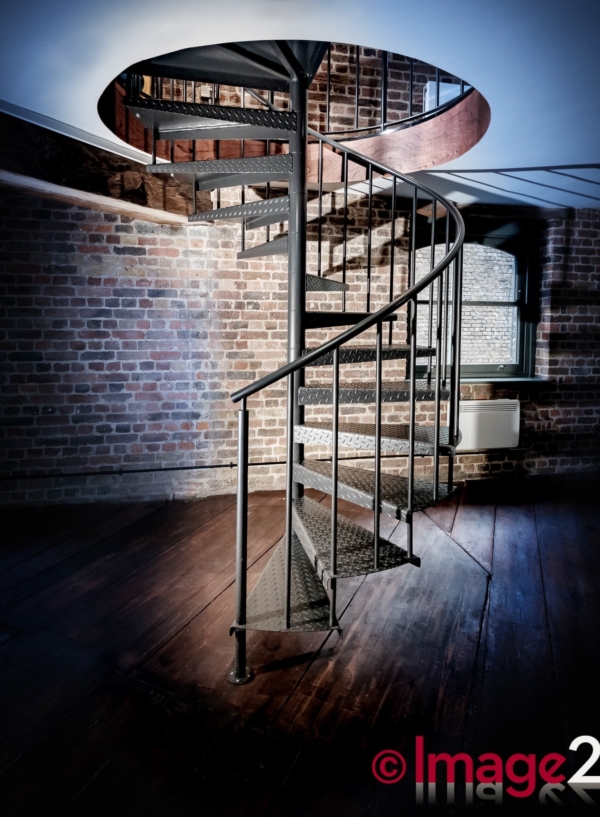 A industrial black metal spiral staircase positioned against a rustic exposed brick wall with timber flooring.