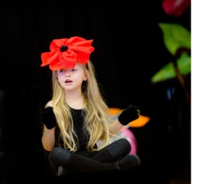 A young girl with long blonde hair wears a large red flower hat and black gloves while performing.