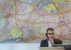 A man in a suit and glasses speaks on the phone in front of a massive, detailed map of West London.