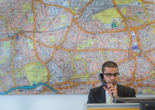 A man in a suit and glasses speaks on the phone in front of a massive, detailed map of West London.