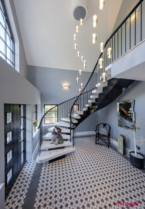 Elegant hallway featuring a curved staircase, patterned tile floor, and a long hanging light fixture with Schnauzer dog sitting on staircase.