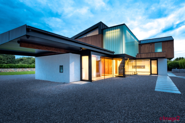 A modern, multi-tiered house at dusk featuring glowing glass sections and a gravel courtyard.