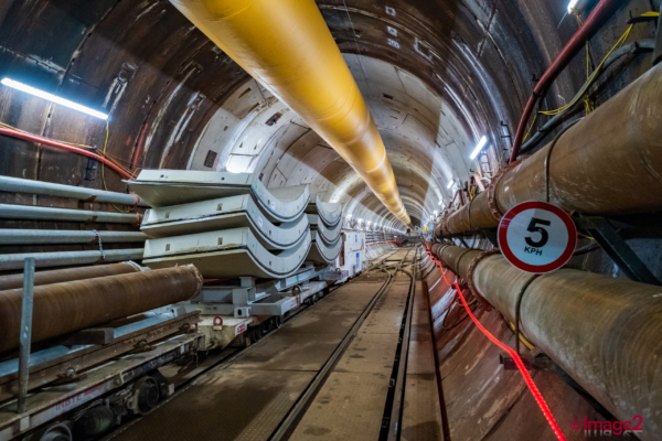 Inside a curved tunnel, flatbed wagons hold concrete segments below a large yellow ventilation pipe.