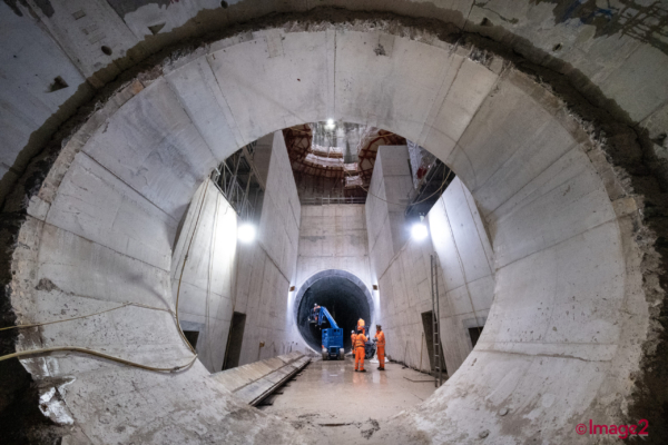 View looking through a circular concrete tunnel opening into a massive shaft where workers stand near a blue cherry picker.