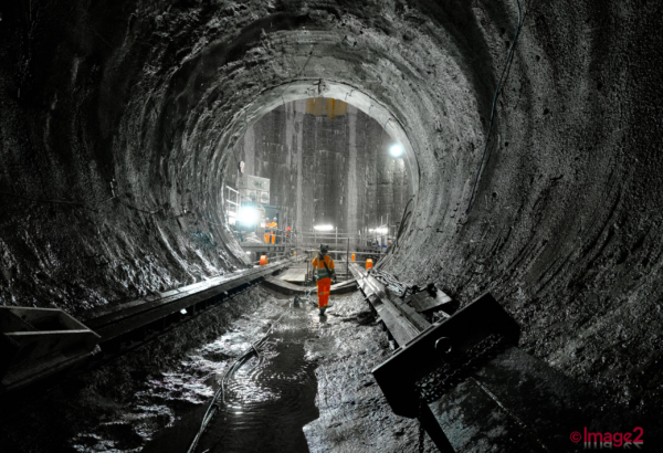 A lone worker in orange safety gear walks away through a rough, wet underground construction tunnel toward a lit opening.