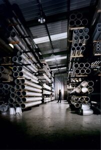 A person stands in a large warehouse aisle surrounded by high racks stacked with industrial metal pipes.