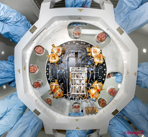 Scientists in blue overalls and hairnets look down into a large, circular piece of machinery.