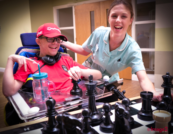 A smiling nurse stands next to a happy man in a wheelchair playing giant chess.