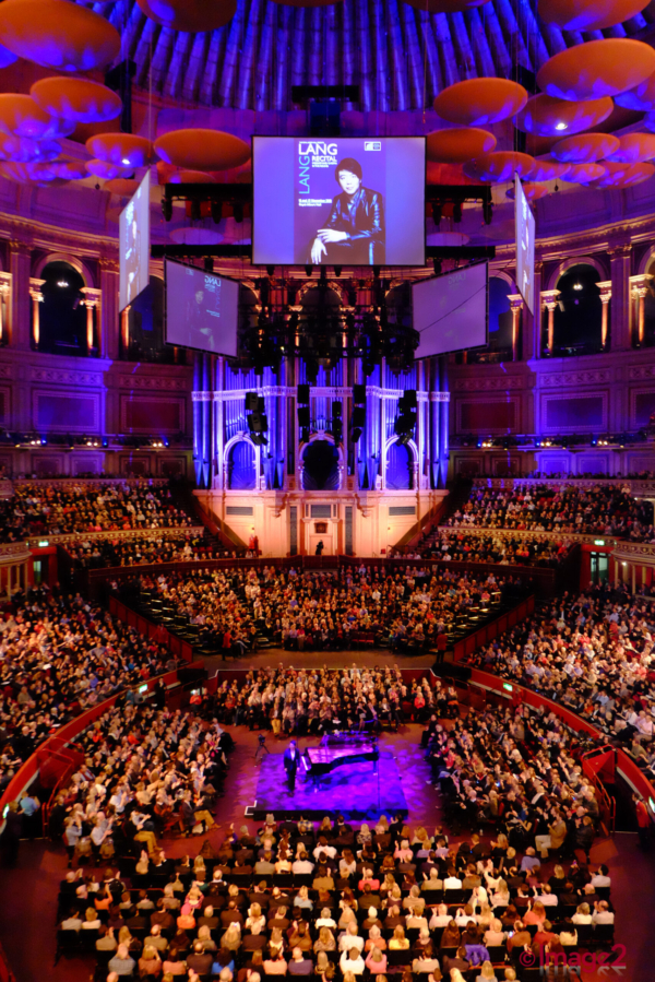 A packed Royal Albert Hall watches a pianist perform on stage under blue lights.