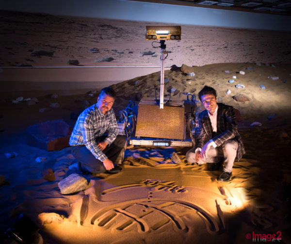 Two men crouch beside a rover on a simulated sandy Mars surface.