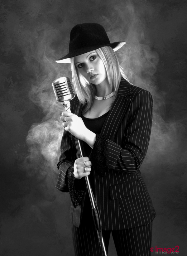 Black and white photo of a blonde woman in a pinstripe suit and trilby hat holding a vintage microphone.