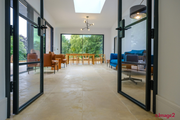 View through open glass doors into a spacious, sunlit living area with a natural stone floor, skylight and large garden window.