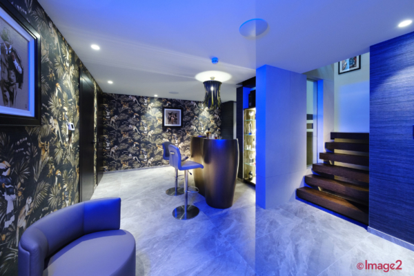Contemporary basement bar with jungle wallpaper, blue lighting, a curved bar counter, and floating stairs.