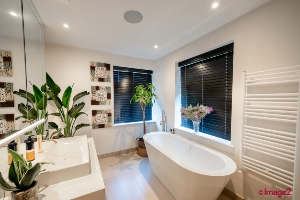 Contemporary bathroom featuring a freestanding tub, black window blinds, a large sink vanity, and leafy plants.