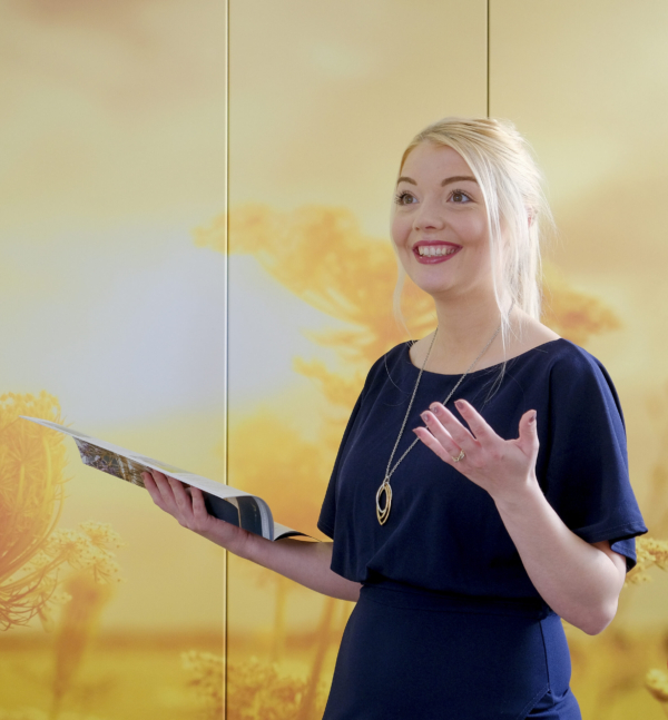 A smiling woman in a dark dress holds an information booklet against a sunny, yellow backdrop.