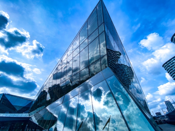 Low-angle shot of a sharp, modern glass building reflecting a bright blue sky filled with dramatic clouds.