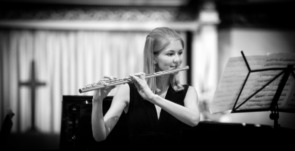 A woman in a black dress plays a silver flute during a performance, captured in a black and white photo.
