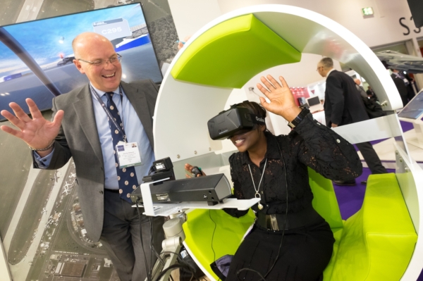 A woman experiences a VR headset in a green pod while a man in a suit laughs and gestures beside her.