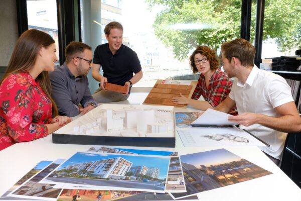 Architects examine sample tiles and building models on a table filled with project images.