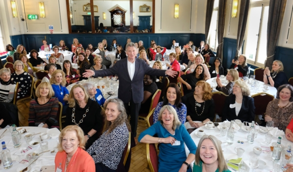A man stands with arms wide in a large hall filled with women seated at round tables for a formal event.