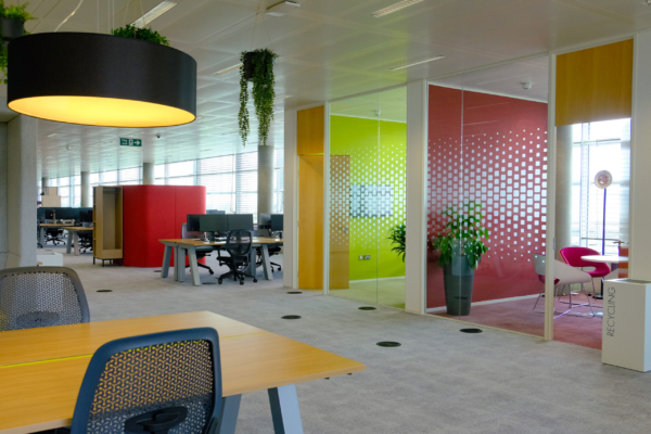 Colourful office space featuring vibrant meeting pods, desks with computers, and hanging plants.