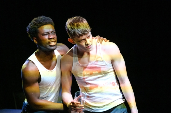 Two distressed men in white Tee shirts tops stand on a dark stage, one holding a glass.