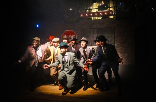 A group of young actors in suits, trilby hats and caps perform on a dimly lit theatre stage.
