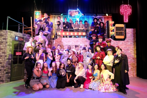 A large theatrical cast poses together in colourful costumes on an elaborate city-themed stage set.