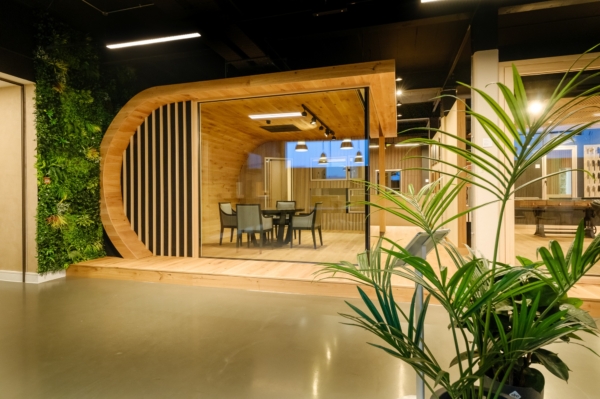 Modern office space featuring a curved wooden meeting pod with glass walls and indoor plants.