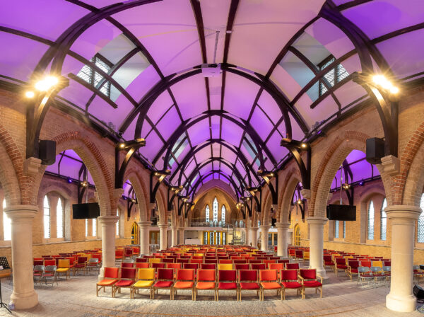 Large hall featuring brick arches, a vaulted wooden ceiling lit in purple, and rows of red and yellow seats.