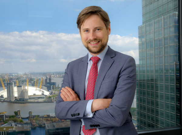 A smiling man in a suit stands with arms crossed against a London city skyline background.