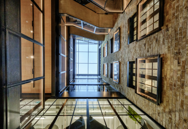 A low-angle look up an industrial atrium with exposed brick, steel mesh walkways, and a glass skylight.