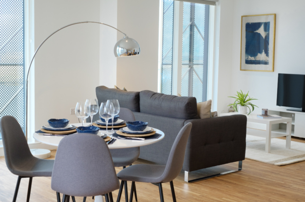 Modern dining area with a round table, grey chairs, an arched floor lamp, and a living area in the background.
