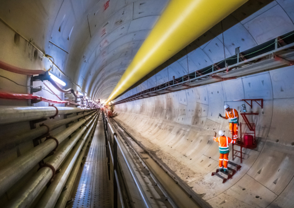 Two workers in high-visibility orange uniforms and hard hats inspect the curved wall of a subterranean tunnel.