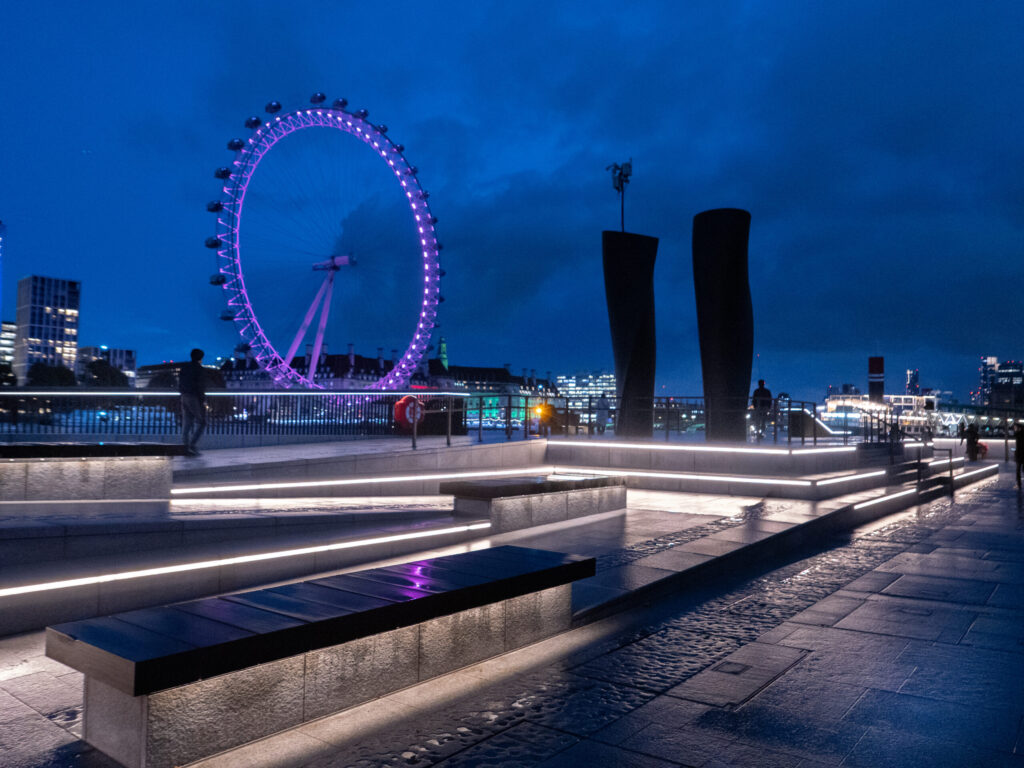 London Eye lit in purple at dusk, viewed from Victoria Embankment with glowing benches and tall sculptures.