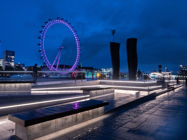 London Eye lit in purple at dusk, viewed from Victoria Embankment with glowing benches and tall sculptures.