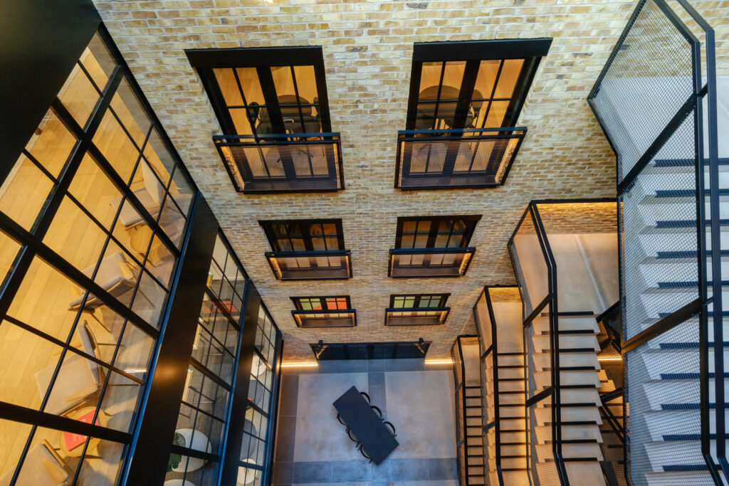A dramatic top-down look into an indoor atrium courtyard, flanked by staircases and glass-walled rooms.