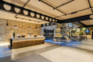 A spacious reception lobby with a wooden "GRAINHOUSE" desk, industrial beams, and a modern staircase.